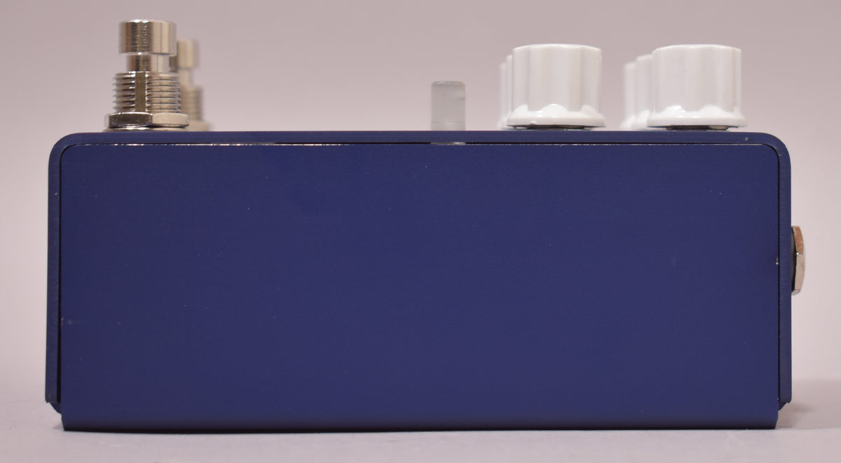 The dark blue pedal is viewed from the side, showing the top edge with silver footswitches and white control knobs.
