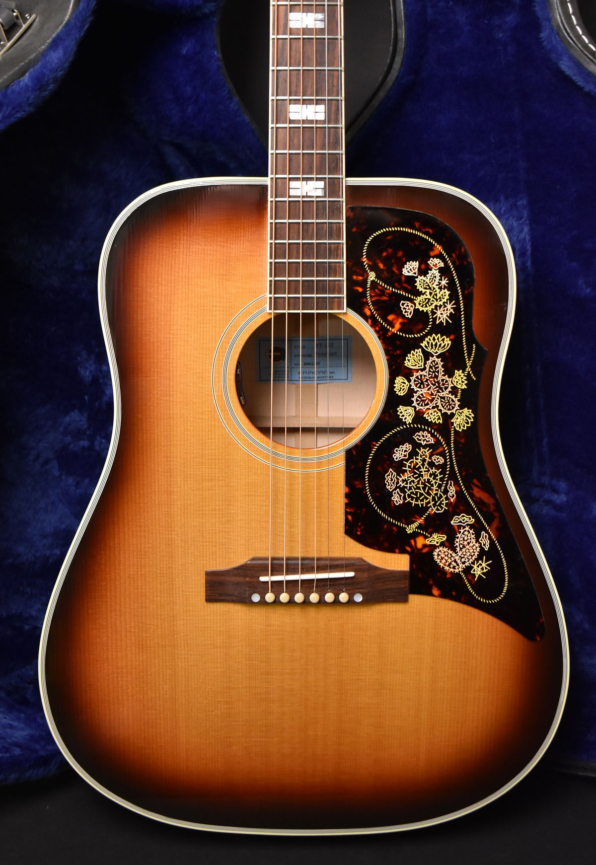 The sunburst acoustic guitar is framed from the neck to the lower bout, showing a tortoiseshell pickguard with floral etching.