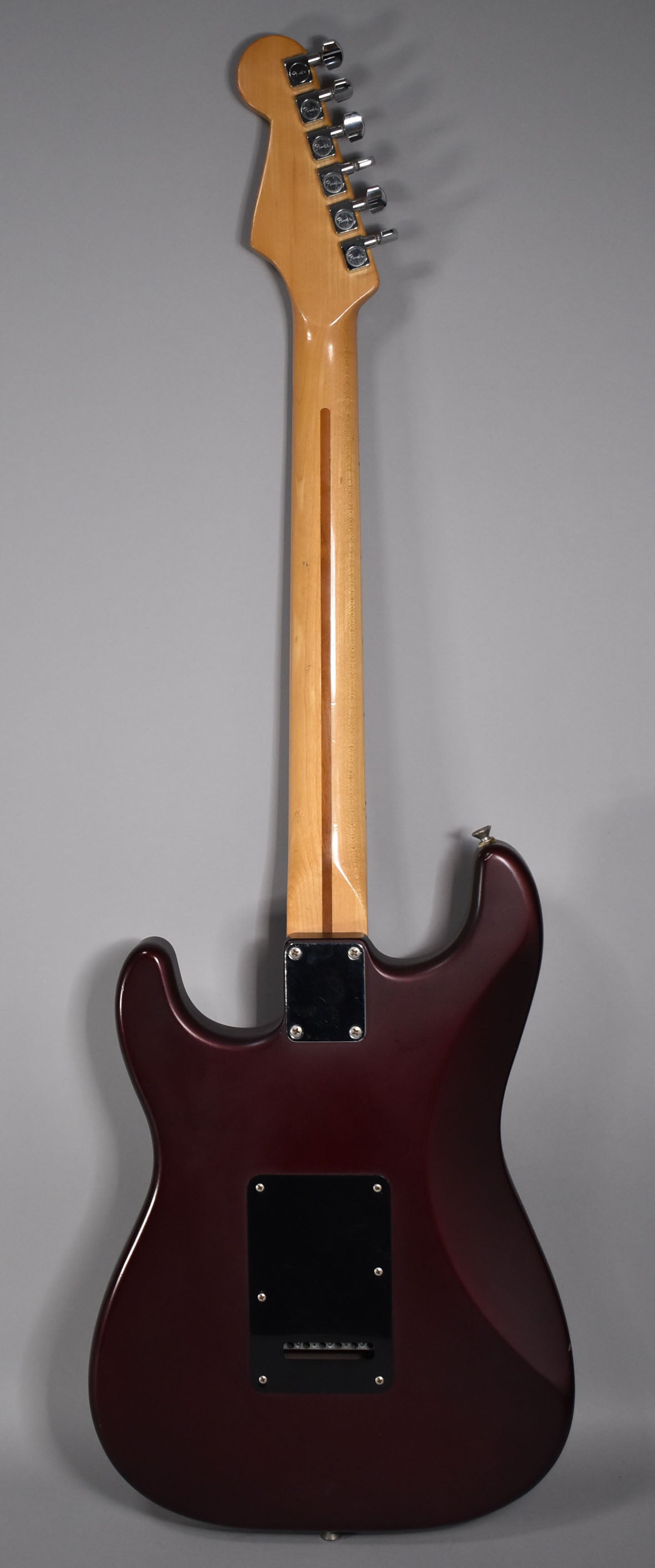 The back of the metallic red Stratocaster shows a maple neck, chrome tuners, a black neck plate, and a black tremolo cavity cover.