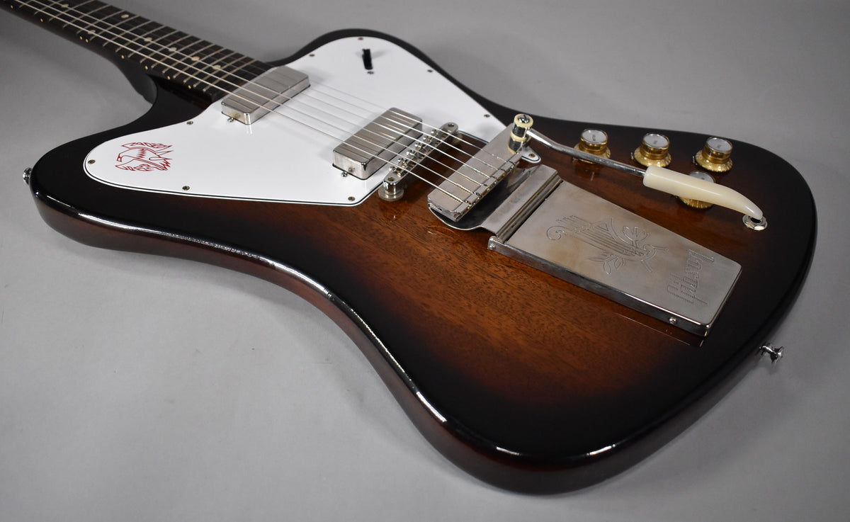 The Firebird's body is angled, showing its dark brown sunburst finish, white pickguard with red bird graphic, and chrome hardware.