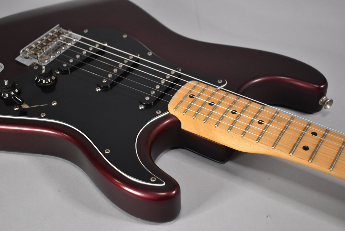 Angled view of a metallic red Stratocaster body with a black pickguard, maple neck, and visible wear around the volume knob.