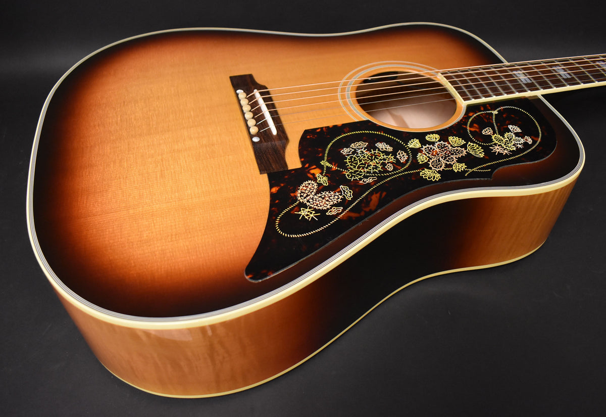 The sunburst acoustic guitar's body is angled, showing the ornate pickguard with gold and white floral patterns, and the bridge.