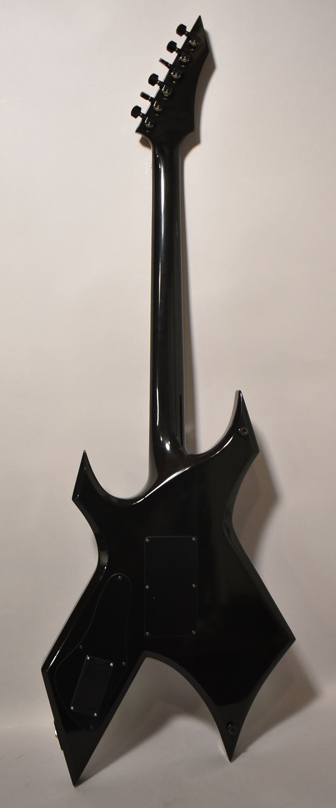 Rear view of a glossy black electric guitar with a pointy headstock, six black tuners, and two control cavity covers.