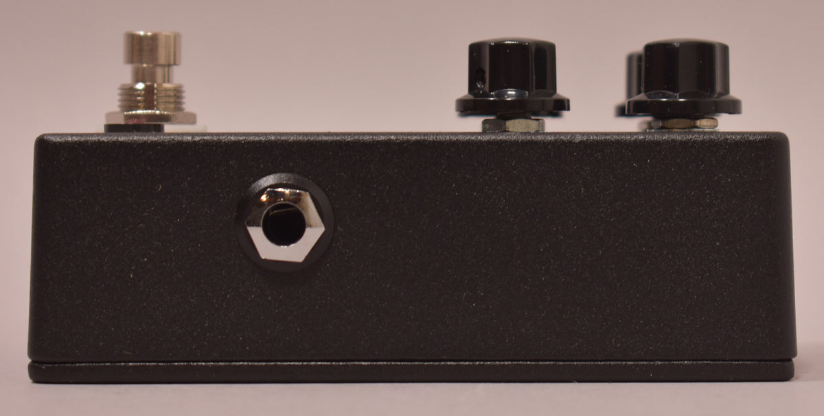The black textured pedal is viewed from the side, showing a silver footswitch, a silver input jack, and two black control knobs.