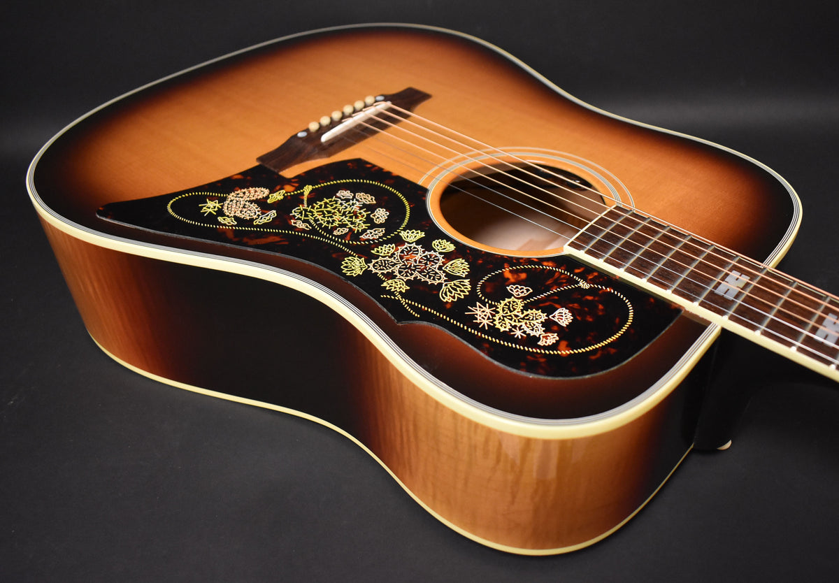 The sunburst acoustic guitar is angled to show its body, with a tortoiseshell pickguard featuring ornate gold and red floral designs.