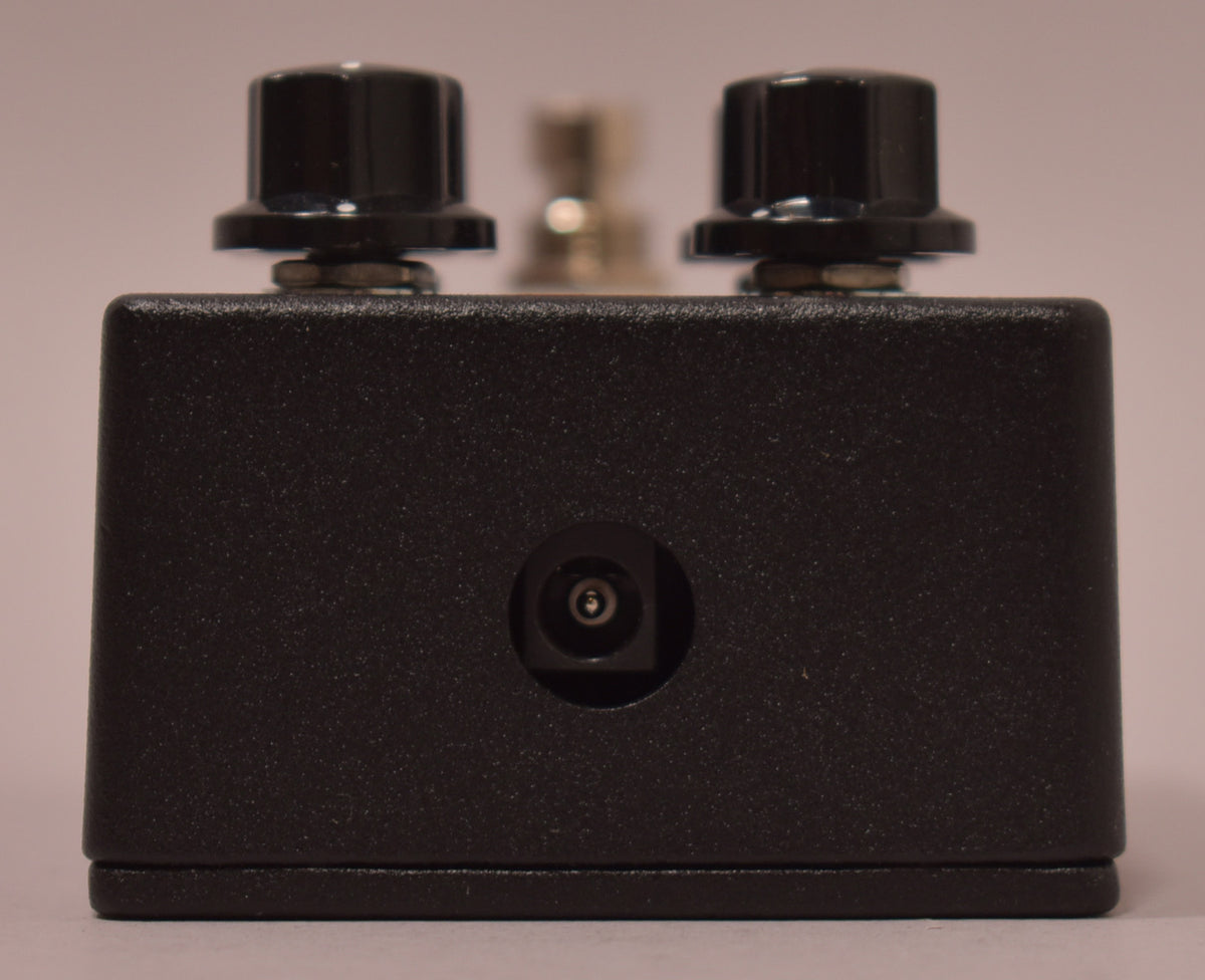 The black textured pedal is viewed from the rear, showing two black control knobs and a centered power input jack.