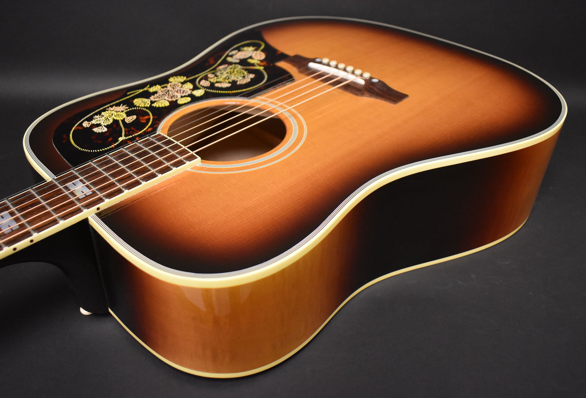 The sunburst acoustic guitar is angled to show its glossy body, with a floral pickguard and white binding around the edges.
