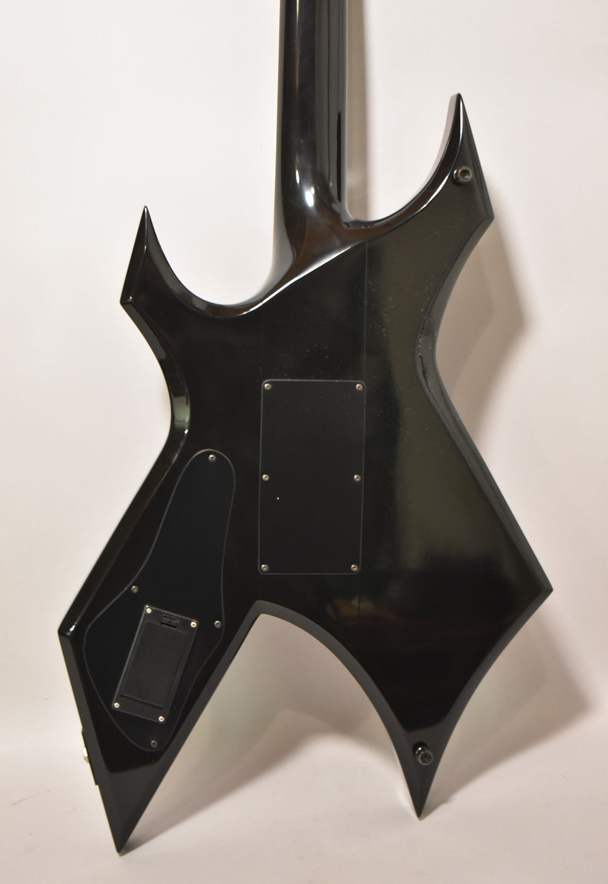 The back of a glossy black Warlock guitar, showing the neck joint, two control cavity covers, and a battery compartment.