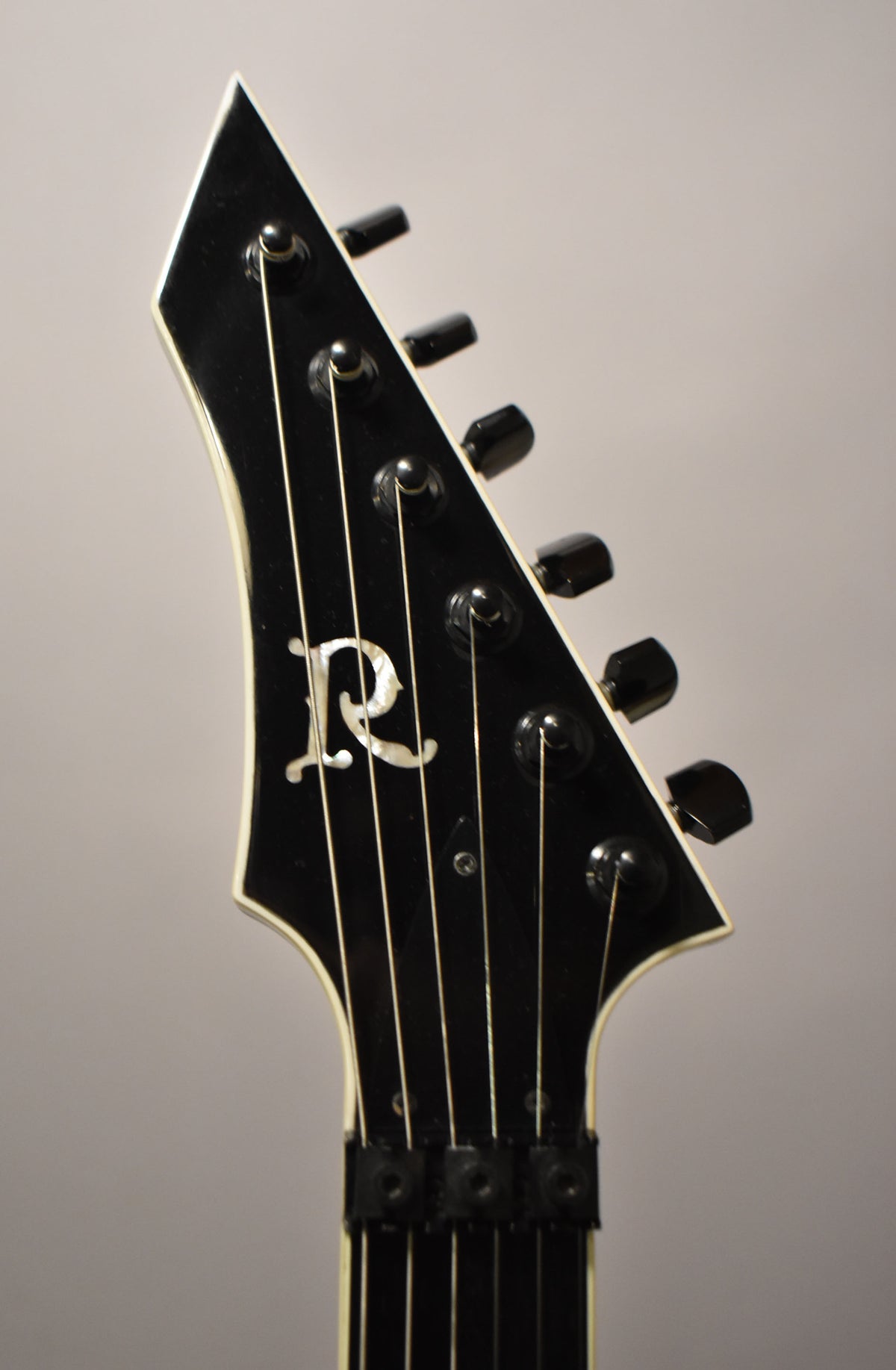 Close-up of the black gloss headstock with white binding, showing the mother-of-pearl "R" inlay, black tuners, and locking nut.