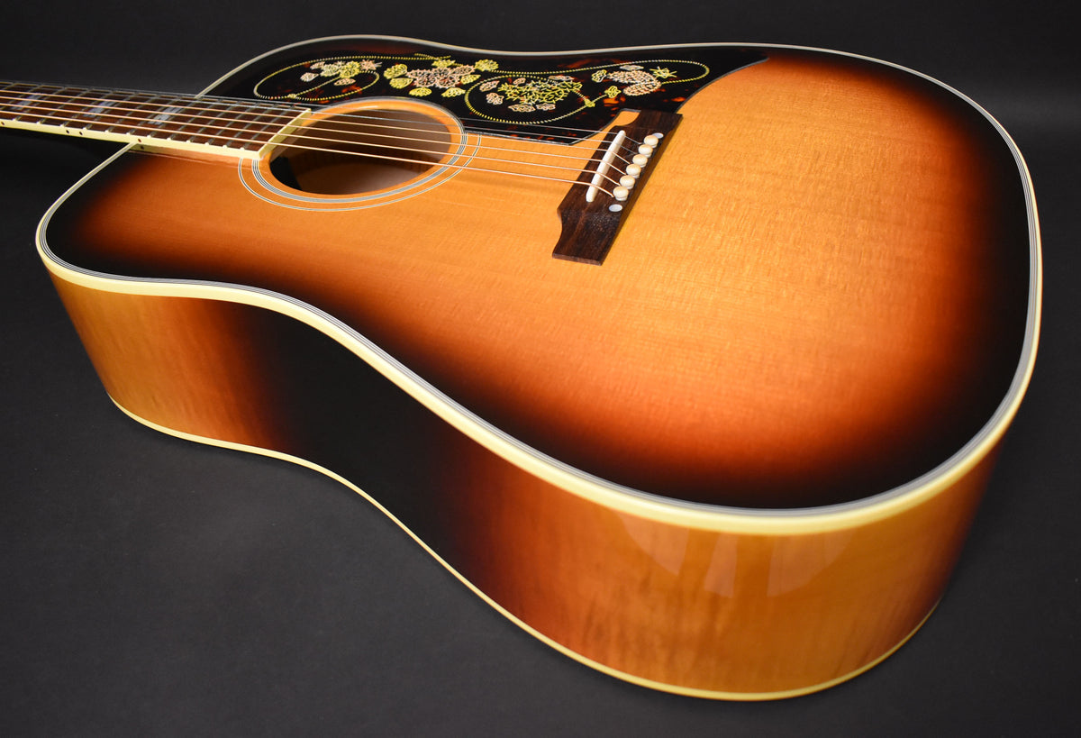 The sunburst acoustic guitar is angled from the upper left, showing its glossy body, pickguard with floral inlay, and bridge.