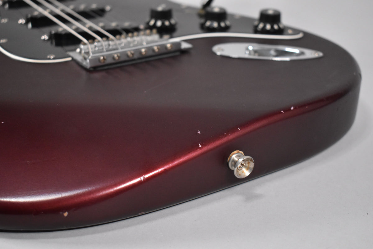 Close-up of the lower body of a metallic red Stratocaster, showing the output jack, strap button, and several chips and scratches on the finish.