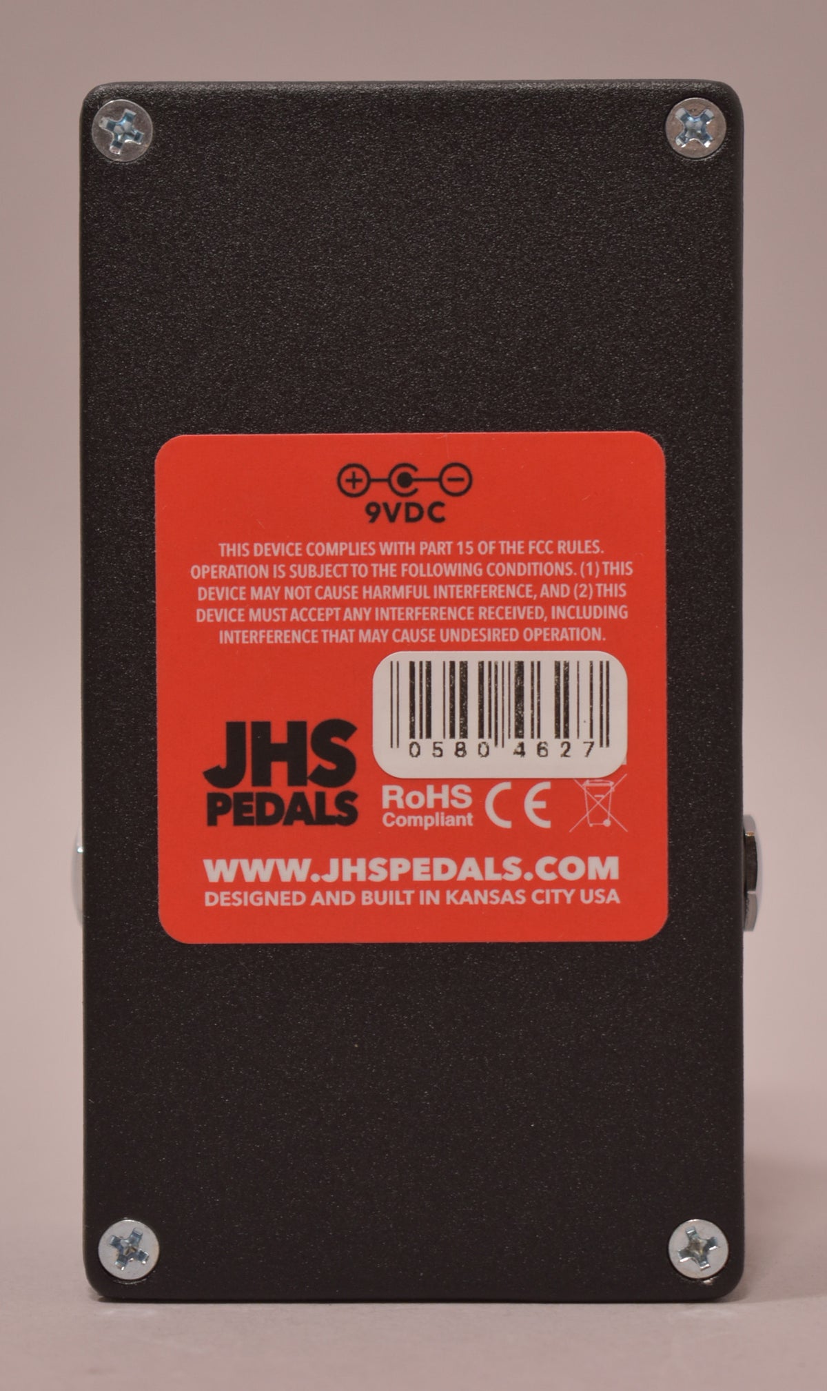 The pedal's textured black underside is visible, featuring a large red sticker with white text and a barcode, secured by four silver screws.