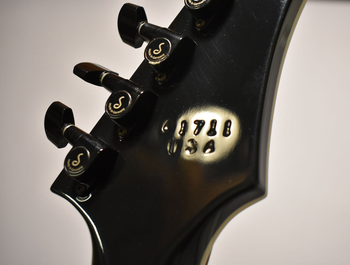Close-up of the glossy black headstock rear, showing three black tuners with gold "S" logos and an embossed serial number.