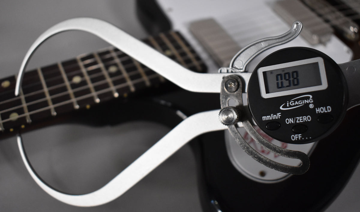 A digital caliper measures the dark fretboard of the guitar, displaying "0.98" on its screen.