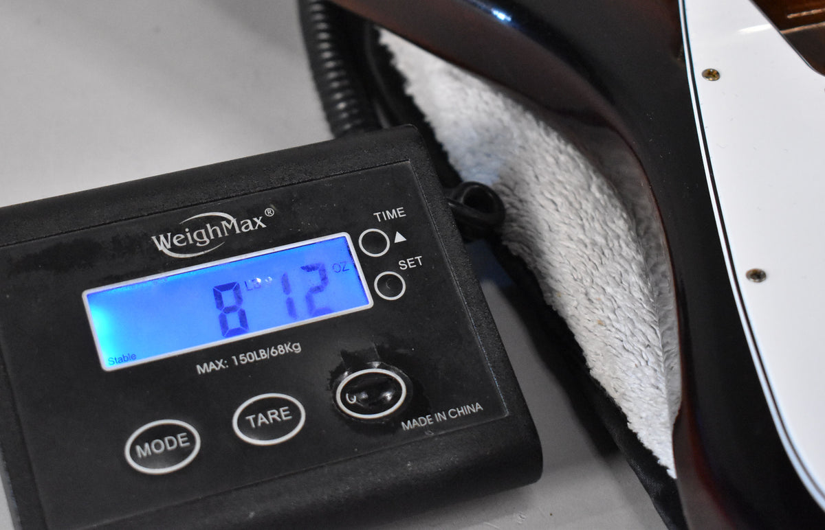 A digital scale displays "8 LB 12 OZ" next to the guitar's dark sunburst body and white pickguard, resting in a plush case.