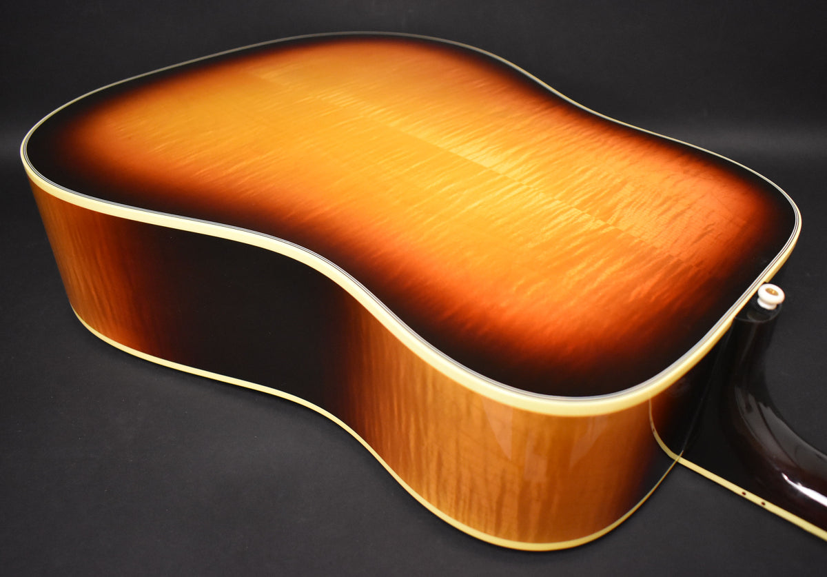The back of a sunburst acoustic guitar, showing the flame maple grain, with a white binding and a strap button visible at the bottom.