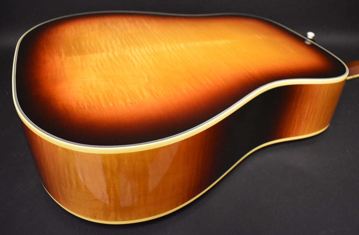 The back of a sunburst acoustic guitar, showing the flame maple grain and cream binding, with a strap button visible at the top.