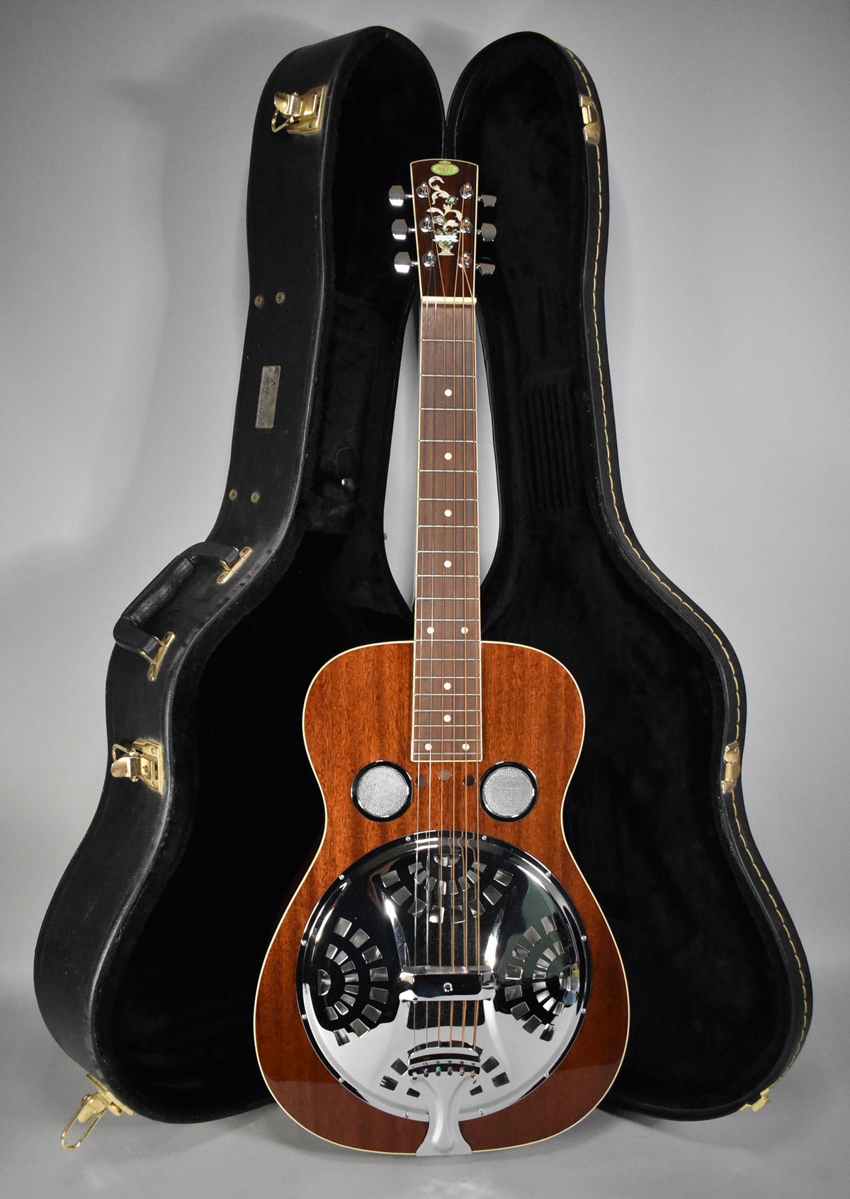 A full-front view of a natural gloss resonator guitar with a chrome cover plate and two sound screens, standing in its open hard case.