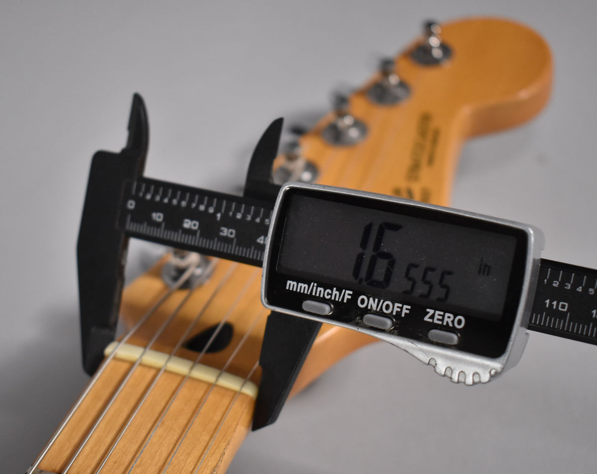 A digital caliper measures the nut width of the maple neck and fretboard at 1.655 inches, with the headstock blurred in the background.
