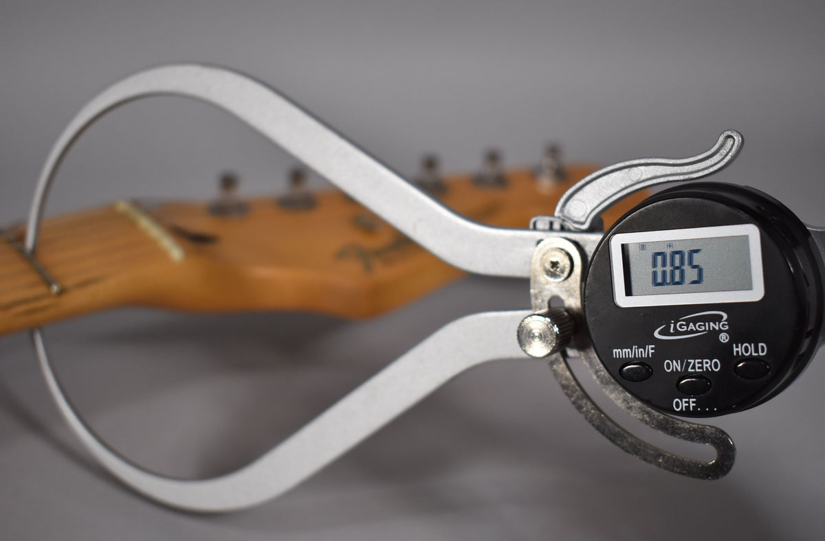 A digital caliper measures the nut width of the guitar's light-colored maple neck, showing a reading of 0.85.