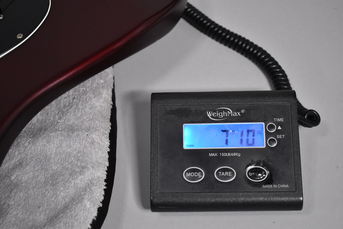 The satin metallic red Stratocaster body rests on a grey cloth, with a digital scale displaying "7 LB 10 OZ" in the foreground.