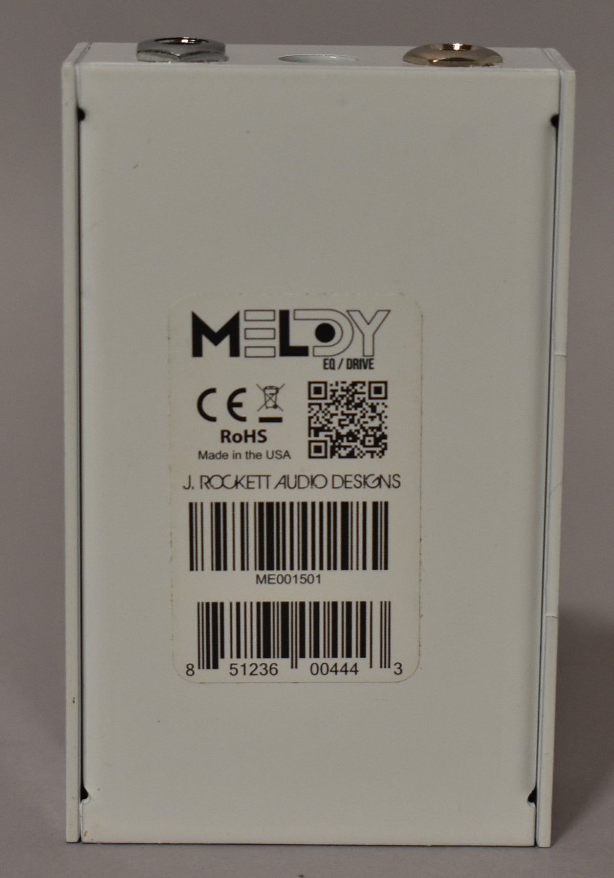 The white pedal's back is shown, featuring a large product label with a barcode and serial number, and two input jacks on top.