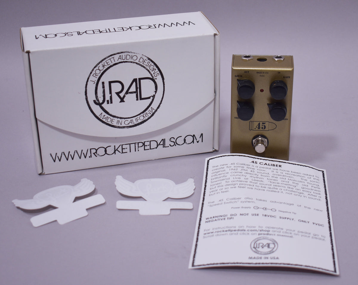 A gold-finished overdrive pedal with four black knobs and a silver footswitch, shown with its white box, manual, and stickers.