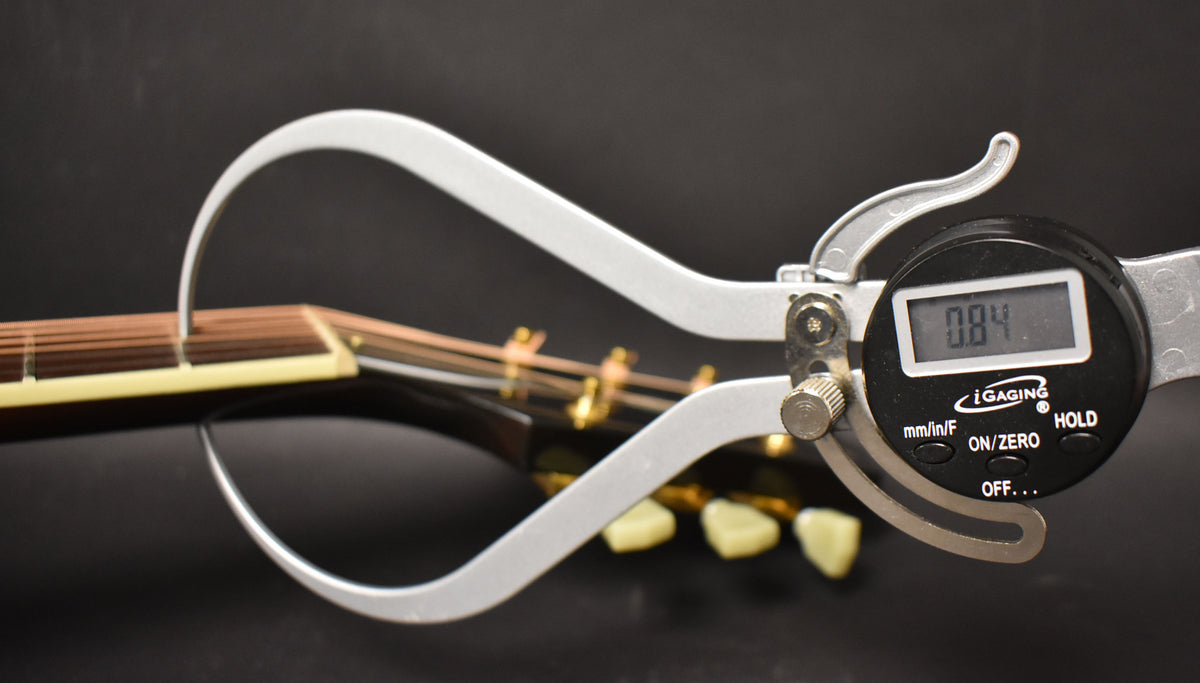 A digital caliper measures the guitar's action at the 12th fret, showing 0.84 on the display, with the headstock blurred behind.