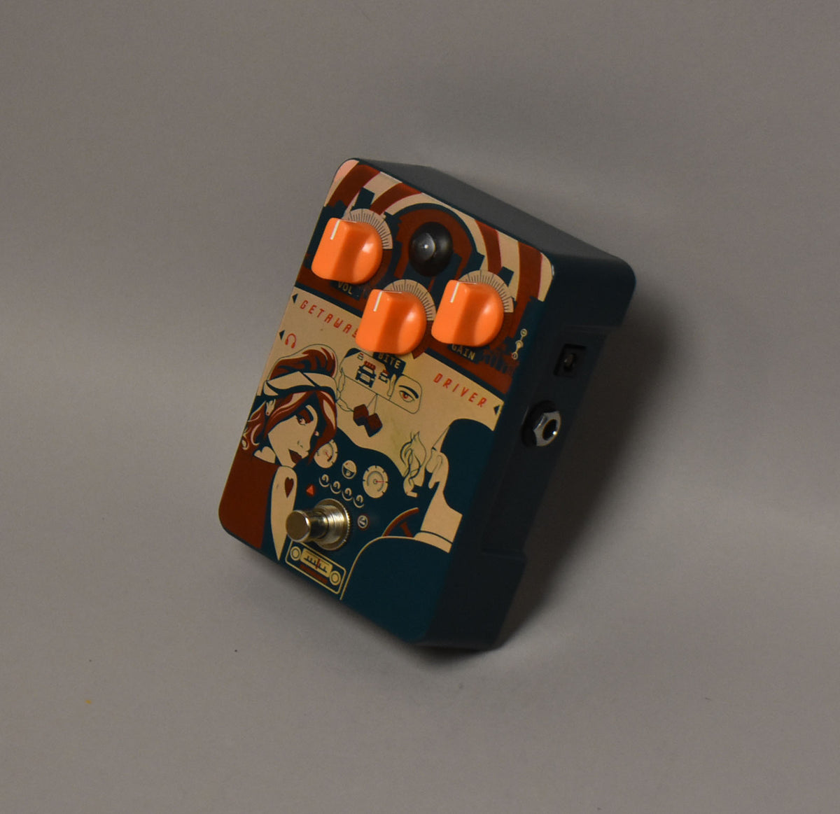 The pedal is angled, showing its top with pop-art graphics, three orange knobs, a footswitch, and the right side with two jacks.