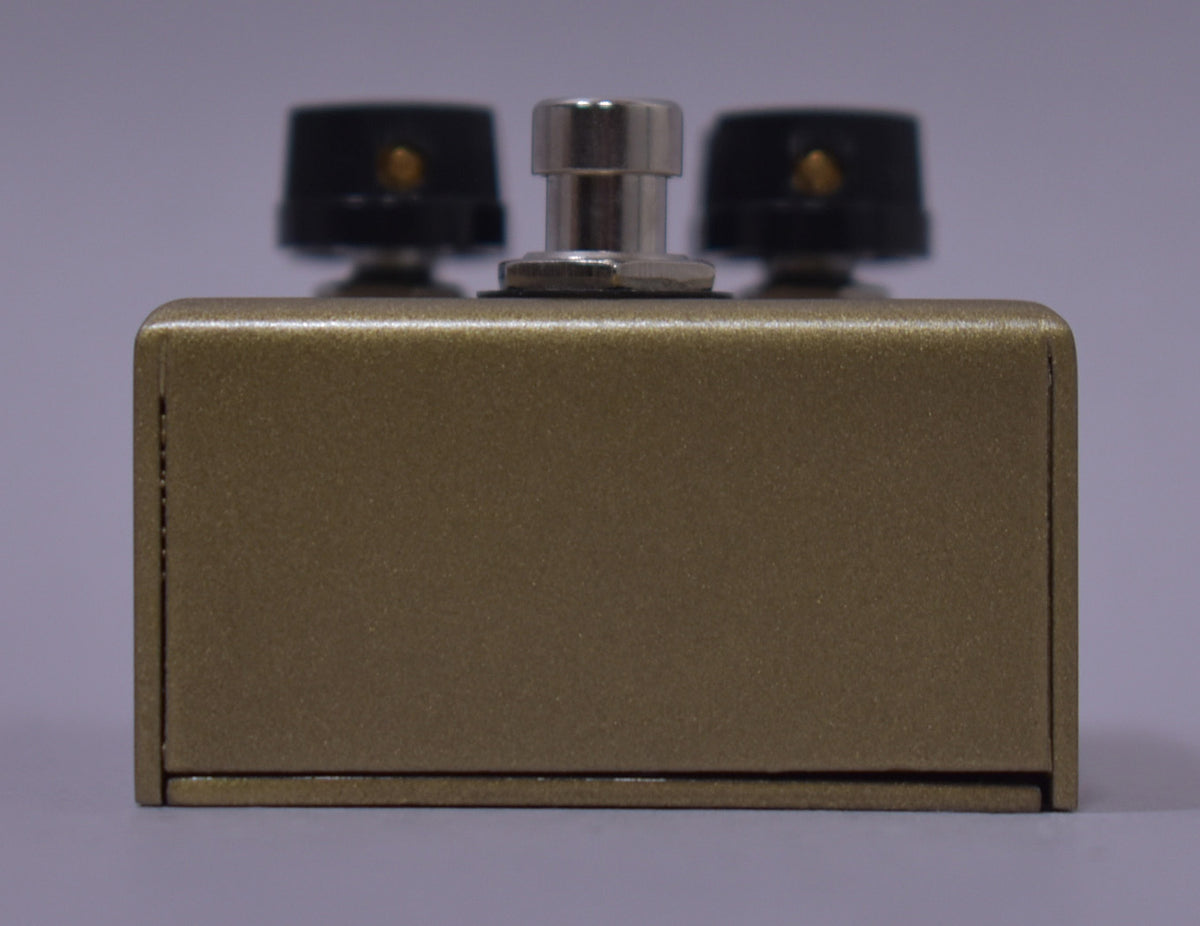 The gold-flecked, textured bronze pedal is viewed from the front, showing the silver footswitch and two black control knobs.