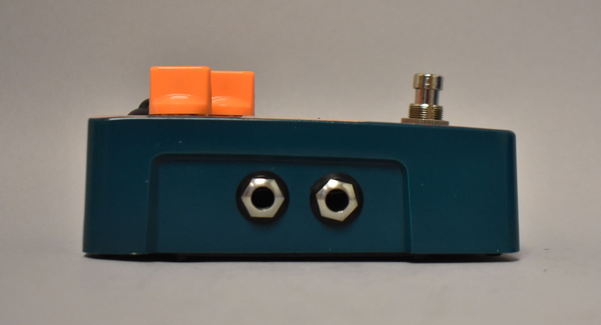 The teal pedal is viewed from the side, showing two input jacks, two orange square knobs, and a silver footswitch.