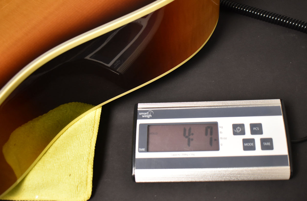 The sunburst acoustic guitar body, with cream binding, rests on a yellow cloth and a digital scale displaying "4.7" lbs.