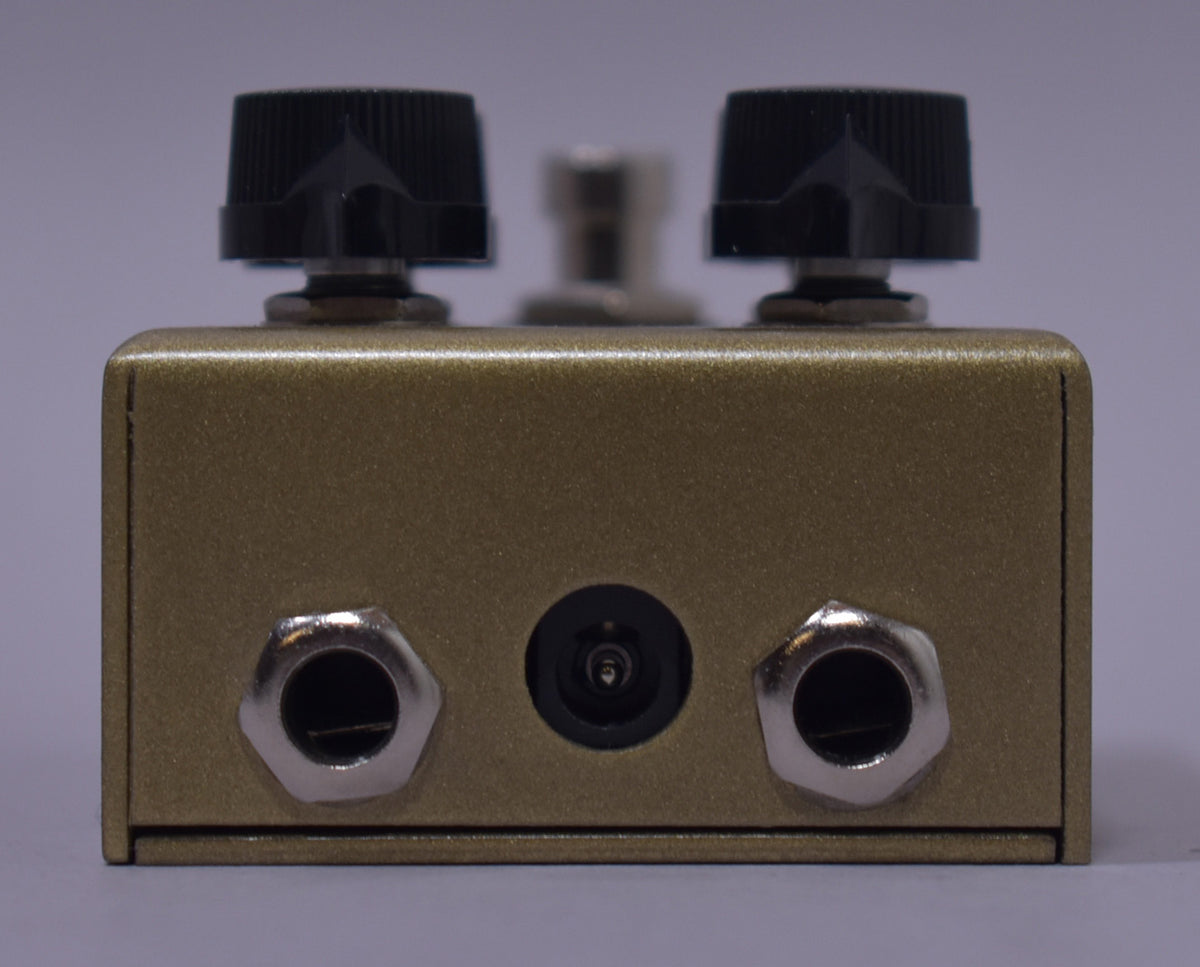 The gold-sparkle pedal is viewed from the back, showing two silver input jacks, a power input, and two black control knobs.