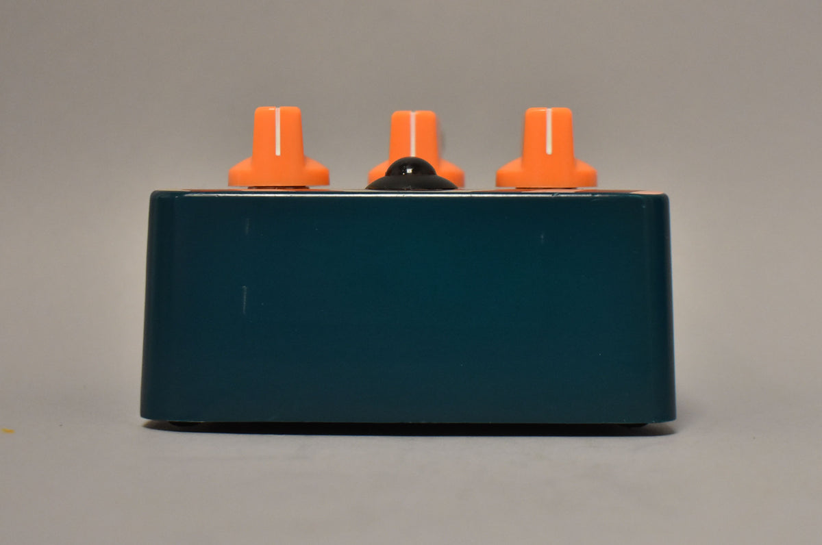 The dark teal pedal is viewed from the back, showing three orange knobs and a black footswitch on top.