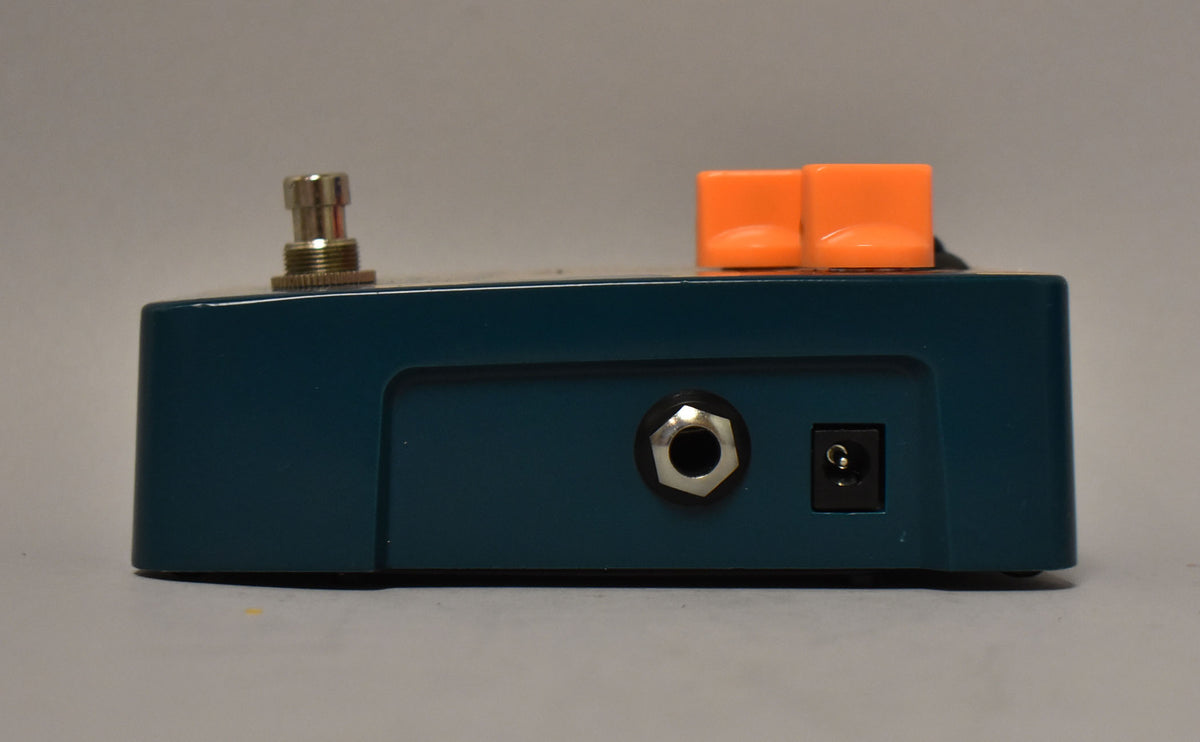 Side view of a dark teal pedal with a silver footswitch, two orange square knobs, a silver input jack, and a black power input.