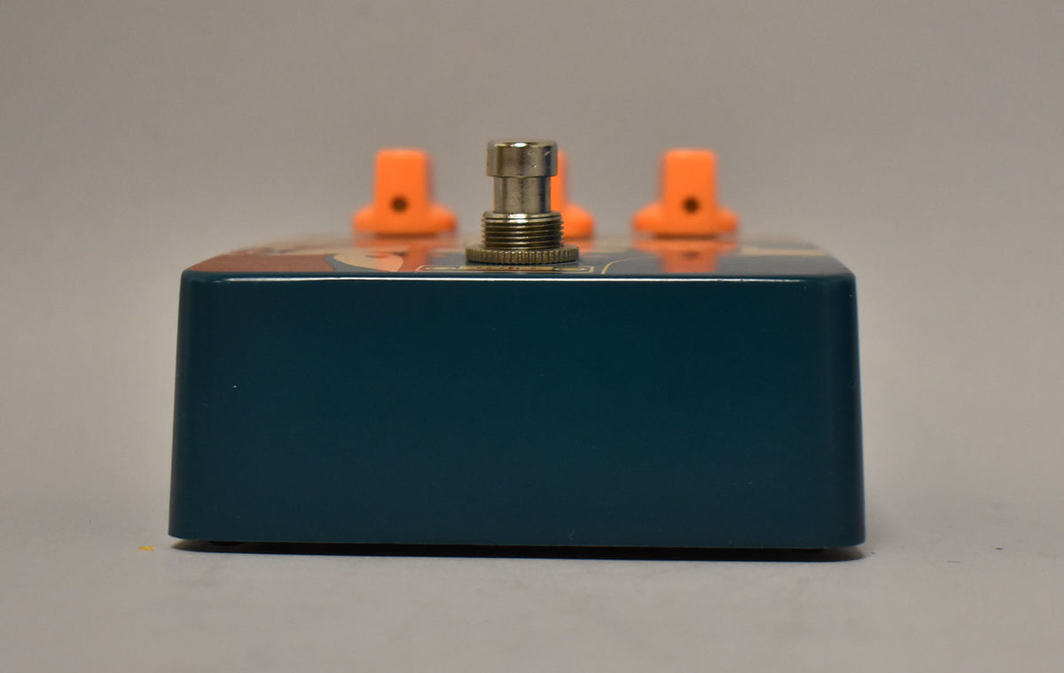 The dark teal pedal is viewed from the side, showing a silver footswitch and three orange control knobs on top.