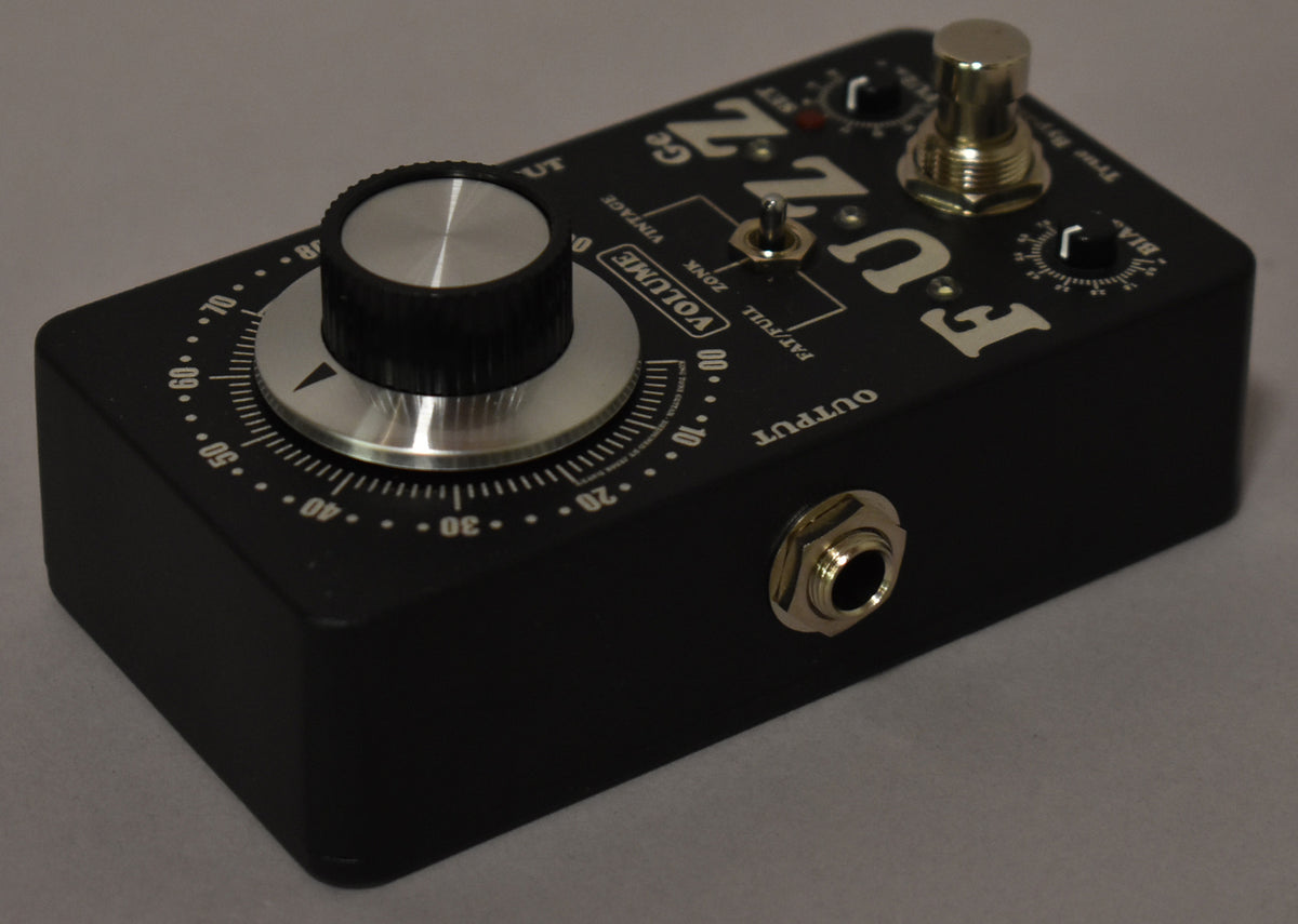 A black effect pedal is angled from the right, showing its top with a large silver volume knob, two smaller black knobs, and a toggle switch.