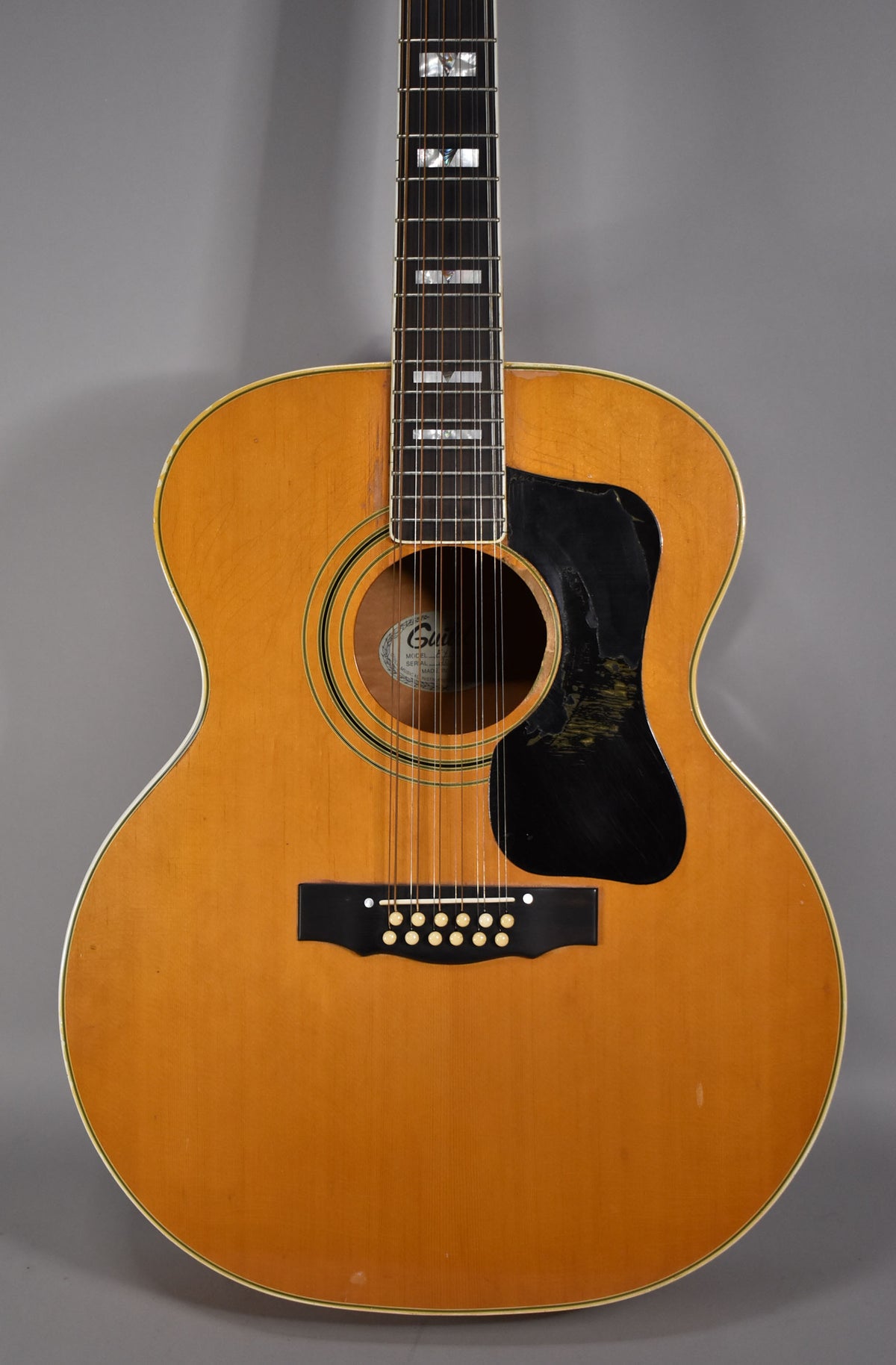 Full front view of a blonde 12-string acoustic guitar with a black pickguard, showing wear and checking on the body and neck.