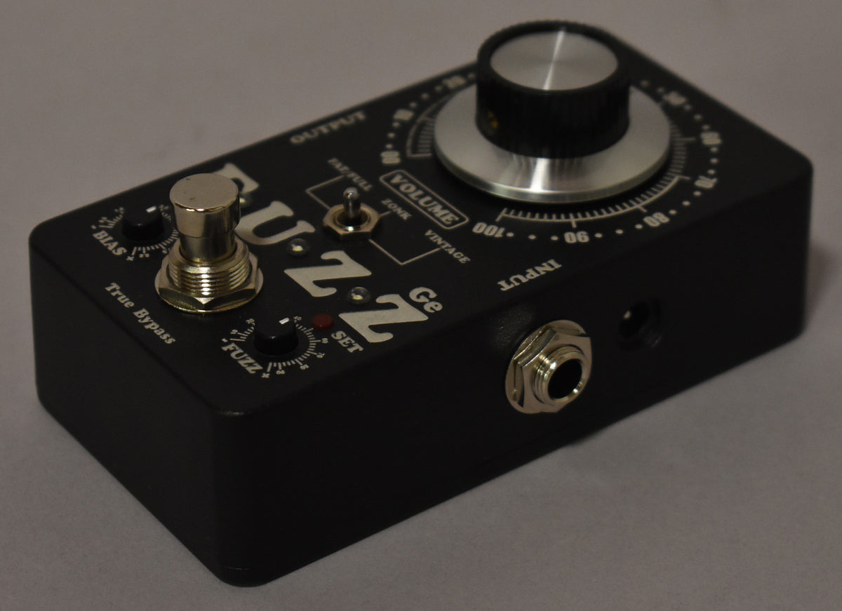 A black guitar pedal is angled from the right, showing its top with silver knobs, a footswitch, and an input jack on the right side.