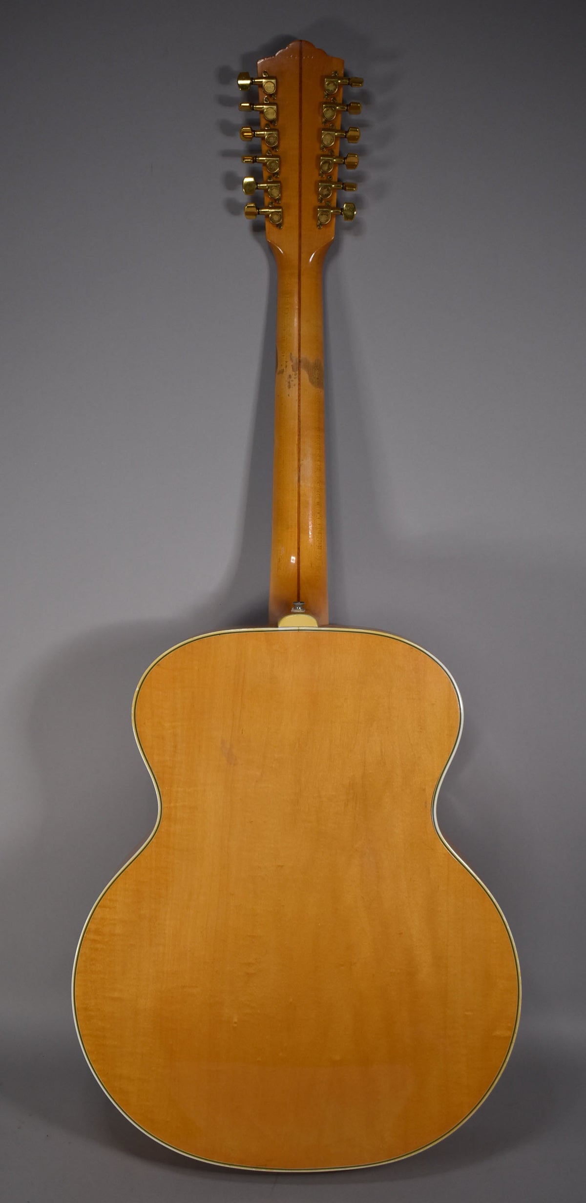The back of a blonde jumbo 12-string acoustic guitar, showing the full body, neck, and headstock with gold tuners.
