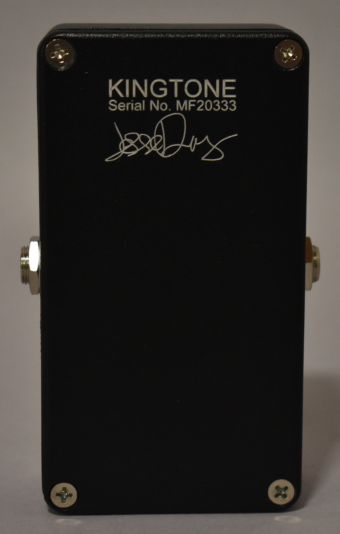 The black effect pedal is viewed from the back, showing the serial number and a white signature, with two silver jacks on the sides.