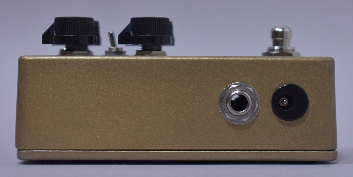 The gold-finished pedal is viewed from the side, showing two black knobs, a toggle switch, a jack, and a power input.