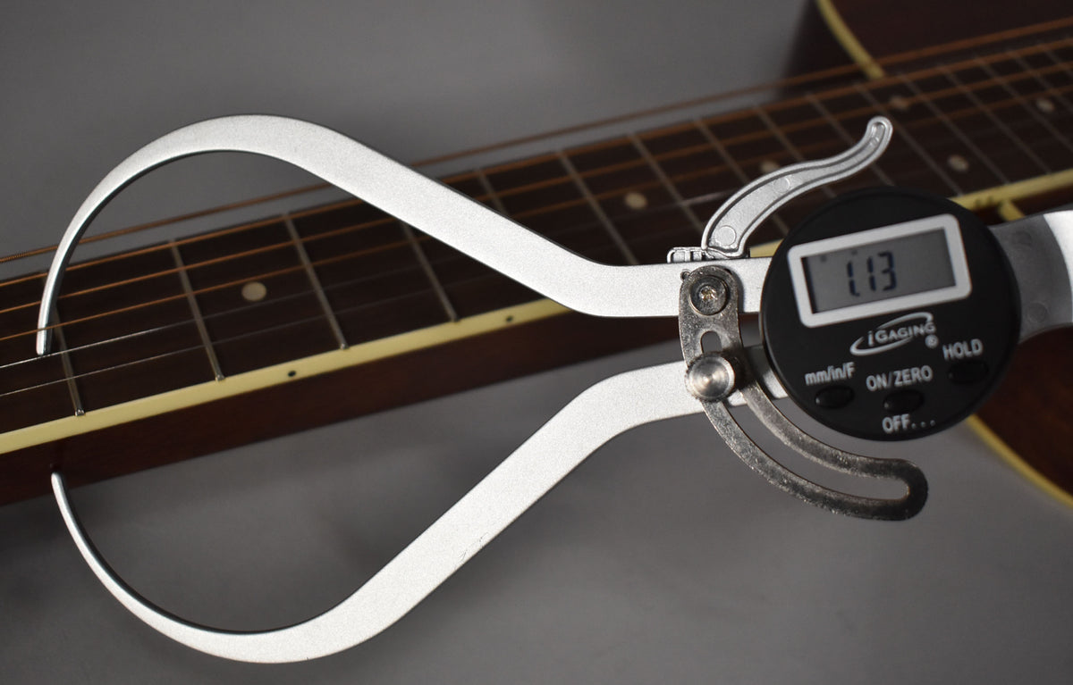 A digital caliper measures the string height at the 12th fret, showing 1.13mm, on the dark brown fretboard with dot inlays.