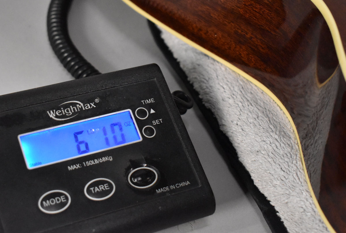 A digital scale displays "6 LB 10 OZ" next to the dark brown, gloss-finished guitar body with cream binding, resting in a grey plush case.