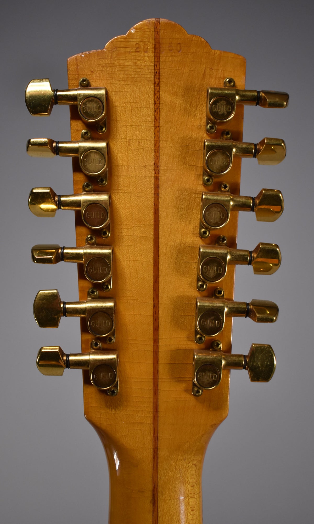 The back of a light blonde headstock with a dark wood stripe down the middle, featuring 12 gold tuning machines.