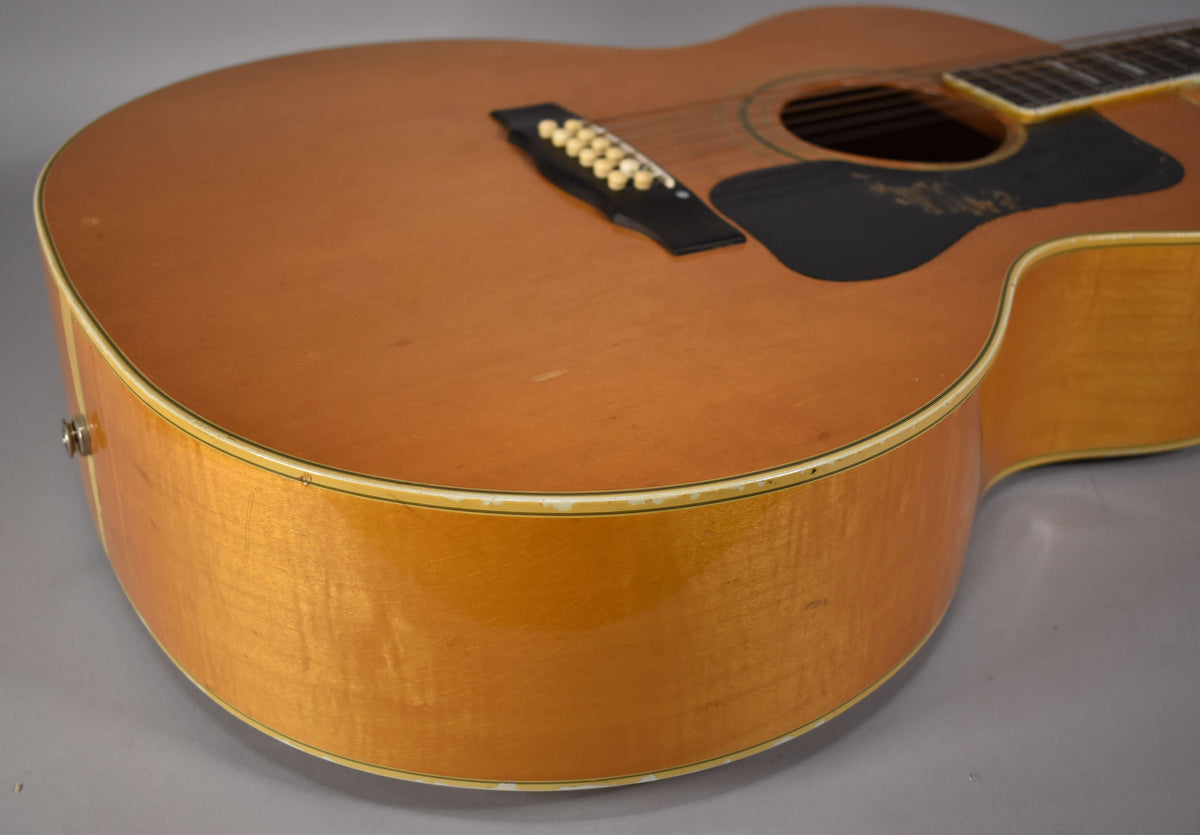 The lower bout of a blonde jumbo acoustic guitar, showing a black pickguard, 12 bridge pins, and wear along the binding.