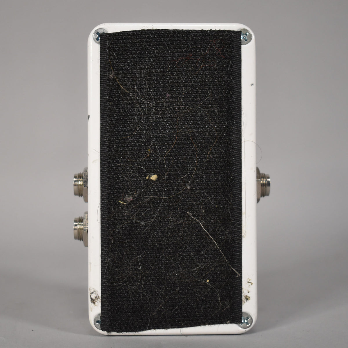 The bottom of a white guitar pedal with a black, fuzzy, lint-covered pad, four screws, and two input jacks on each side.