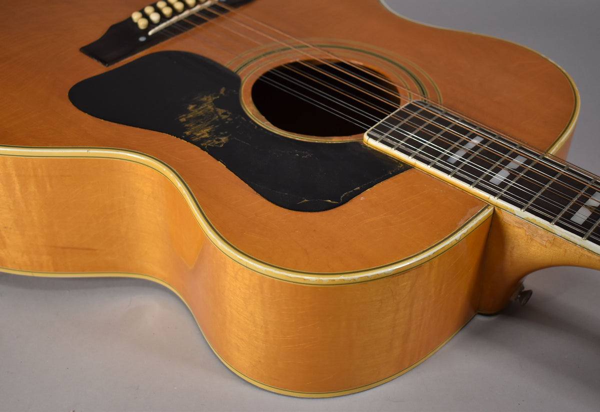 Close-up of the blonde jumbo 12-string acoustic guitar's body, showing a worn black pickguard and white binding with green purfling.