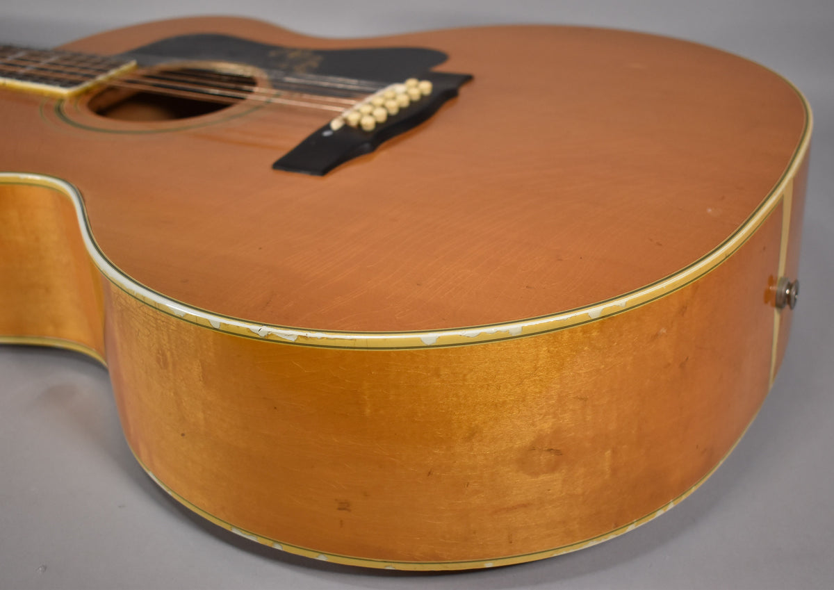 The lower bout of a blonde acoustic guitar, showing a black pickguard, bridge, and worn binding with chipped finish.