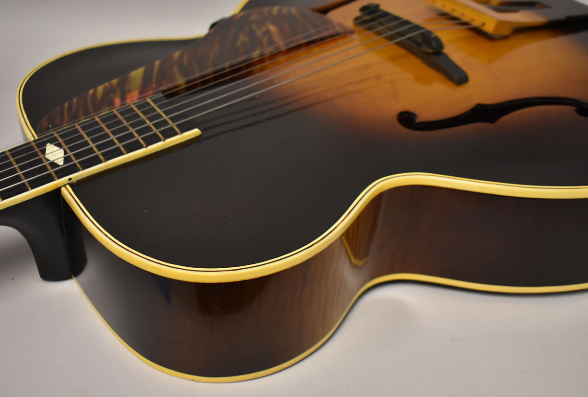 The sunburst archtop's lower bout and side are visible, showing cream binding, a tortoise pickguard, and diamond fretboard inlay.